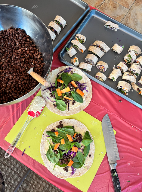 A whole wheat tortilla topped with pieces of carrots, beets, shredded cabbage, spinach, and black beans is placed on a cutting board next to a bowl of drained black beans.